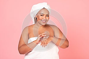 Skin care cosmetics concept. Excited oversized black lady applying moisturizer cream on hands, standing wrapped in towel