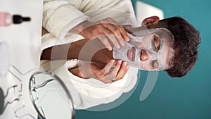 Skin care. Handsome arabic man applying sheet facial mask at his face and looking at himself in front of the mirror