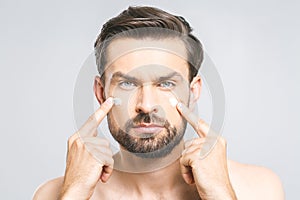 Skin care. Handsome happy young shirtless man applying cream at his face and looking at camera while standing over gray background