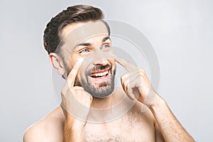 Skin care. Handsome happy young shirtless man applying cream at his face and looking up with smile while standing over gray