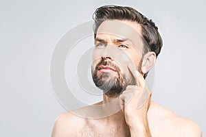 Skin care. Handsome happy young shirtless man applying cream at his face and looking up while standing over gray background and