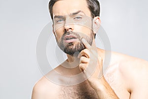 Skin care. Handsome happy young shirtless man applying cream at his face and looking up while standing over gray background and