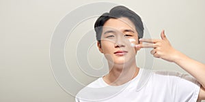 Skin care. Handsome young man applying cream at his face and looking at himself with smile while standing over white background