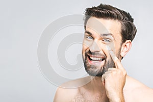 Skin care. Handsome young shirtless man applying cream at his face and looking at camera with smile while standing over gray
