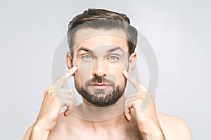 Skin care. Handsome young shirtless man applying cream at his face and looking at camera while standing over gray background and