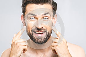 Skin care. Handsome young shirtless man applying cream at his face and looking at himself with smile while standing over gray