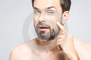 Skin care. Handsome young shirtless man applying cream at his face and looking at himself with smile while standing over gray