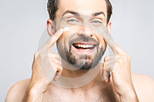 Skin care. Handsome young shirtless man applying cream at his face and looking at himself while standing over gray background and