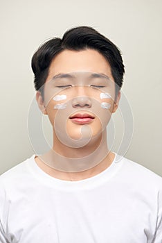 Skin care. Portrait of handsome young man applying cream to his face, isolated on white background