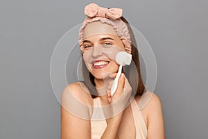 Skin care routine. Morning beauty treatment. Smiling attractive young woman with hair band using face brush for exfoliation posing