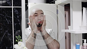 Skin care. Young Caucasian bald man applying facial lotion on his bearded face and looking at mirror. Home male beauty