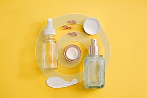 Skin rejuvenation concept - glass face serum bottle, eye cream, face oil capsules on yellow background