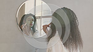 Skin treatment at home. Young african american lady applying natural clay mask on her face, looking at mirror