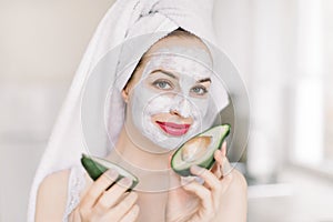 Skin treatment and spa procedures at home. Young pretty woman with a smile holding avocado peaces, with face clay