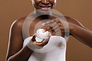 Skin Treatments. Smiling Black Lady Holding Open Jar With Moisturising Cream, Closeup