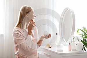 Skincare Concept. Attractive Middle Aged Lady Applying Moisturising Face Cream At Home