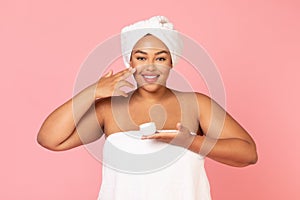 Daily skincare concept. Portrait of overweight african american lady holding cream jar and applying moisturizer on cheek