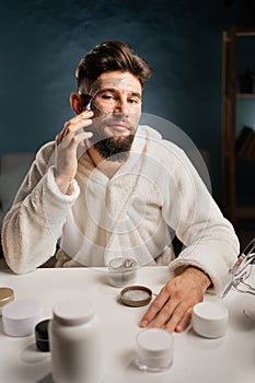 Skincare and cosmetology concept. Bearded man in bathrobe at home spa makes beauty facial procedures. Handsome man take