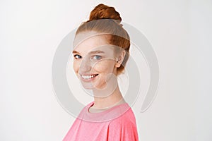Skincare and makeup concept. Close-up of pretty redhead woman with messy bun, blue eyes and freckles, smiling with white