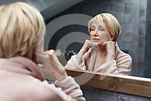 Skincare Routine. Mature Woman Touches Face Skin In Front Of The Mirror Reflection.