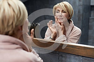 Skincare Routine. Mature Woman Touches Face Skin In Front Of The Mirror Reflection.