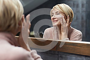 Skincare Routine. Mature Woman Uses Cosmetic Cream On Face Skin In Front Of The Mirror Reflection.