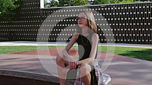 A skinny 12-year-old girl with glasses and a skateboard is sitting on a rollerdrome. Teen sports.