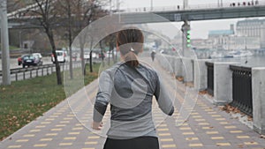 Skinny fit young woman with beautiful body fast running on city quay along river with bridge on background. Female athlete prepari