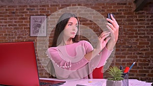Skinny young model swiping her phone and taking selfies with serious look in front of her smartphone on red background