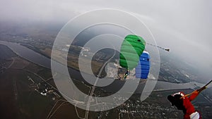 Skydivers with colorful parachutes fly over green field in cloudy sky. Extreme