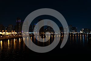 Skyscrapers are reflecting in water. Beautiful view to Sharjah city center lights skyline at night, United Arab Emirates