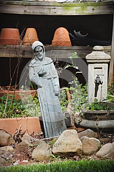 Slanted gray stone St. Francis statue with rocks, plants, fountain, and wood shelf of terra cotta plant pots in background