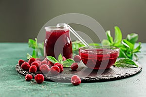 Slate plate with glass jar and bowl of tasty raspberry jam on green table