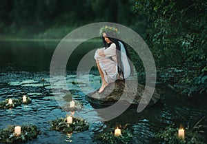 Slavic girl sits on stone on shore lake. Nymph fantasy woman hugs knees. Long black hair. Wreaths of grass, flowers