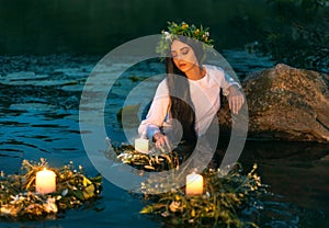 Slavic woman nymph stands in water herbal wreath floa candles burning. Fantasy girl mermaid. White long wet dress