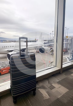 A sleek suitcase stands before a large airport window, offering a view of an aircraft and the expansive tarmac. It's