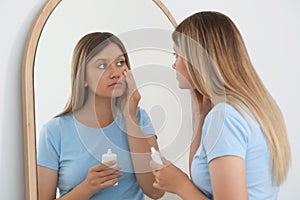 Sleep deprived young woman covering up dark circles with concealer near mirror at home