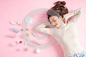 Sleeping Asian woman lying on pink background with her cosmetics. Top view