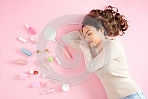 Sleeping Asian woman lying on pink background with her cosmetics. Top view