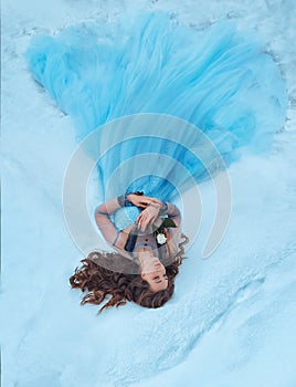 A sleeping beauty lies on the snow with a white rose in her hands. She is dressed in a luxurious, lush, blue dress