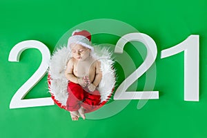 A sleeping child in a Santa costume lies with a candy in his hands on a green isolated background with the numbers 2021, waiting