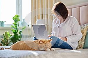 Sleeping ginger cat at home on bed, woman using laptop on background