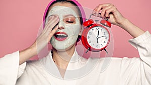 Sleeping girl with mask on face holds alarm clock in hand. Beautiful girl with cosmetic mask on face. Woman wearing face