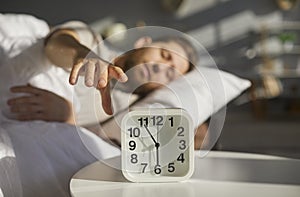 Sleeping man lying on the bed in his bedroom and stopping alarm clock on bedside table at home.