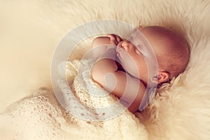 Sleeping newborn baby lying on white fur carpet covered with cream knitted blanket.