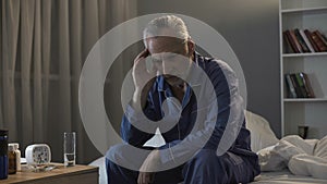 Sleepless senior man suffering from headache, sitting on sofa at night time