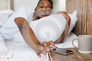 Sleepy Black Man Reaching For Smartphone Turning Off Alarm-Clock Indoor