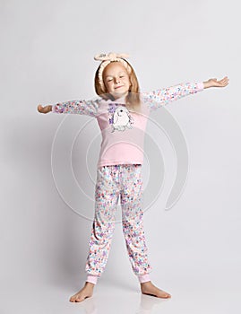 Sleepy blonde kid girl in headband, stylish shirt and pants pyjamas with flower print pattern yawning and stretching
