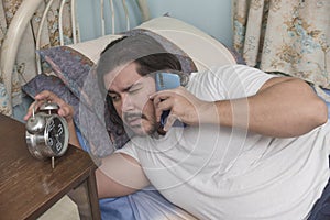 A sleepy and groggy man lying in bed checks the time on his alarm clock while talking to a client. Freelancer taking up rush work photo