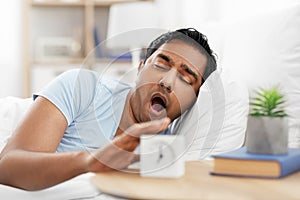 Sleepy indian man with alarm clock in bed yawing